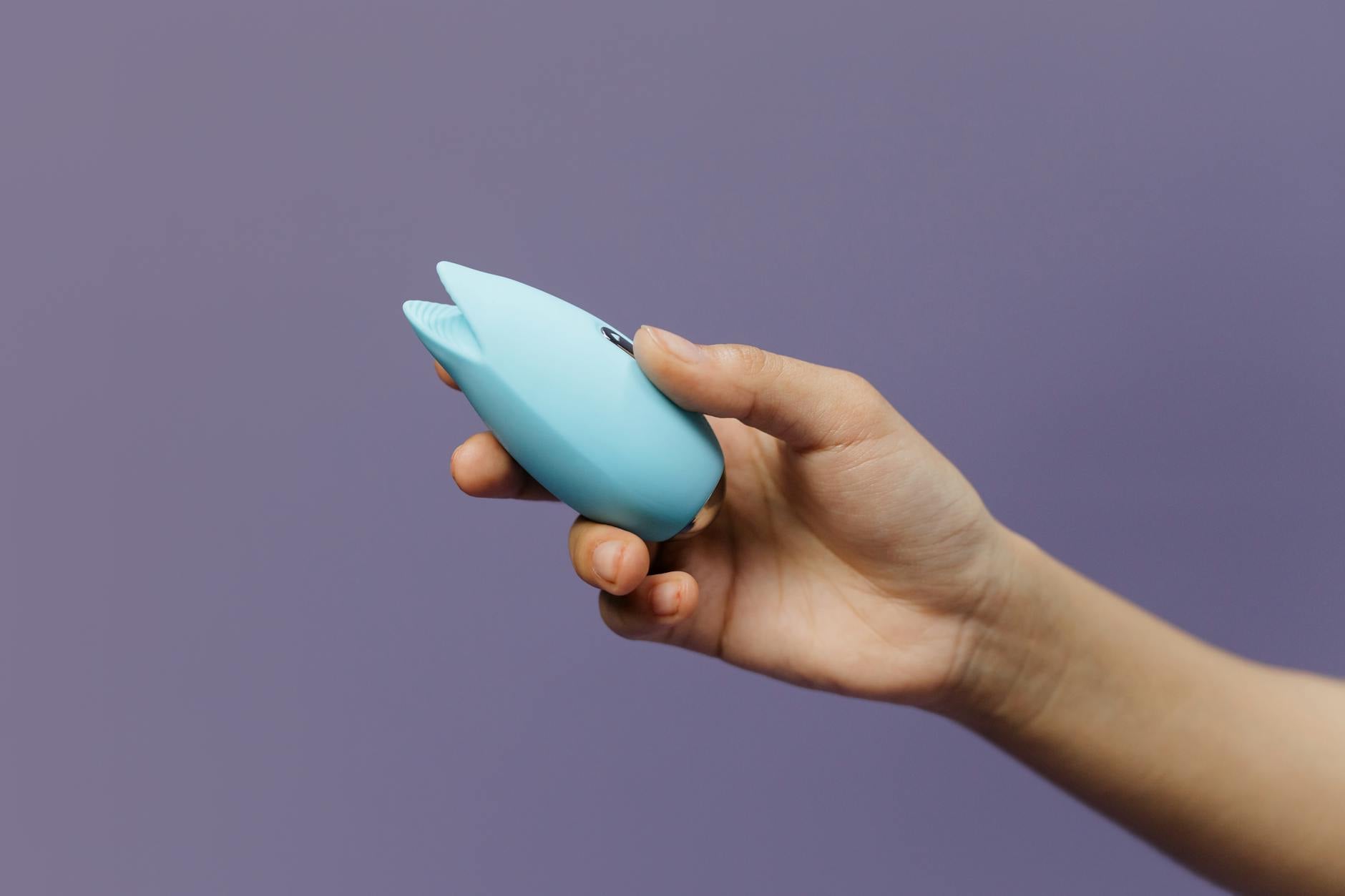 A blue silicone clitoral vibrator held in hand against a solid purple background, promoting self-love and sexuality